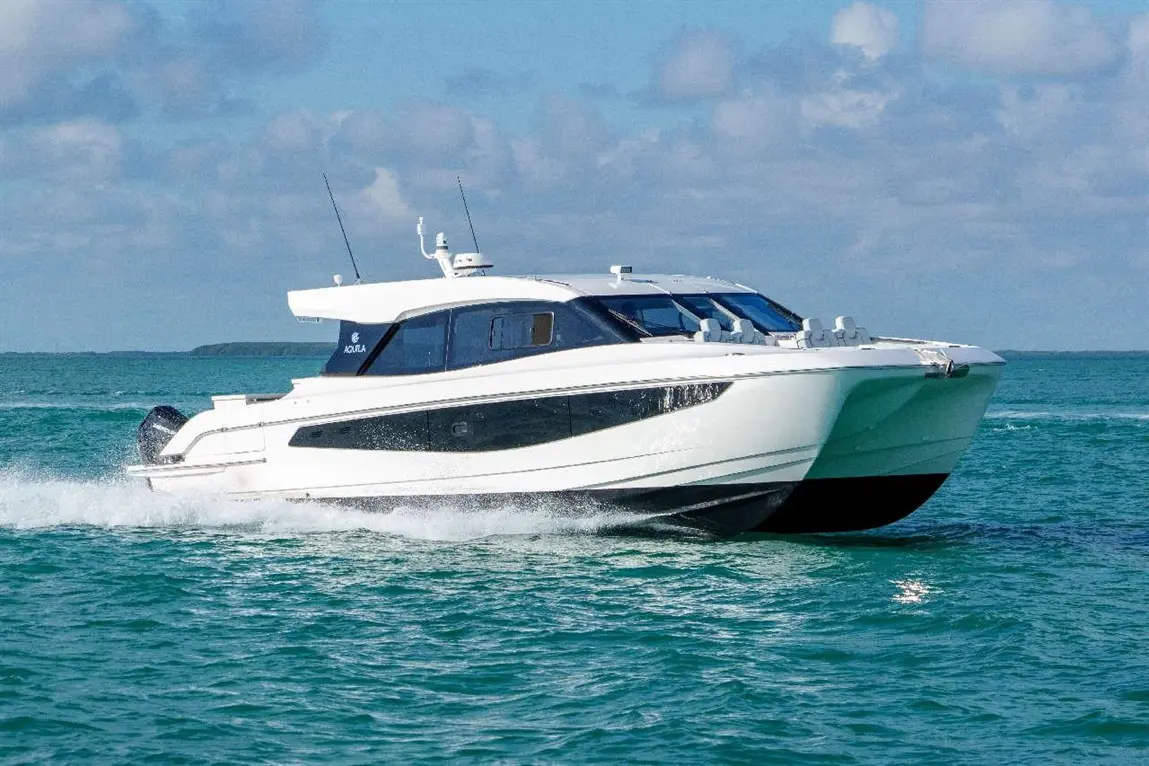 A sleek white boat with a black trim glides through turquoise waters under a partly cloudy sky, creating waves behind it.